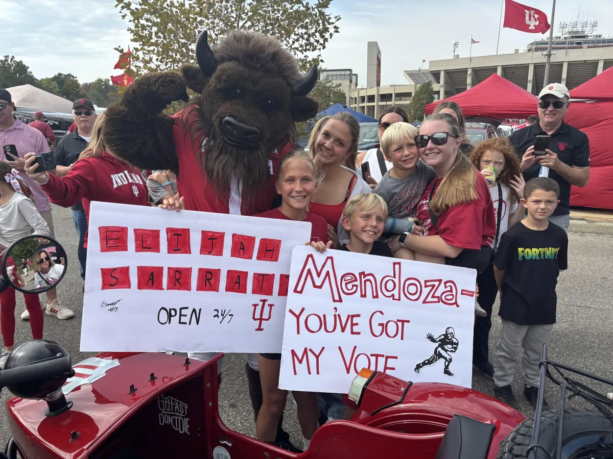Family posing with the IU mascot at a Hoosiers football tailgate, holding fan signs for players Elijah Sarratt and Mendoza outside Memorial Stadium in Bloomington, Indiana