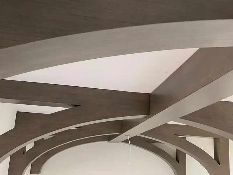 Masterful finish carpentry showcasing intricate geometric curved and straight grey wood-grain beams forming decorative arches within white panels on a custom modern ceiling, meticulously installed by Brothers Home Group in Naples, FL.
