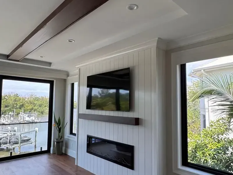 A luxurious modern living room feature wall in Naples, Florida, with a custom white vertical shiplap paneling wall, built-in linear electric fireplace, dark wood floating mantel, and mounted TV. Above is a detailed coffered ceiling with dark accent beams, crafted by Brothers Home Group