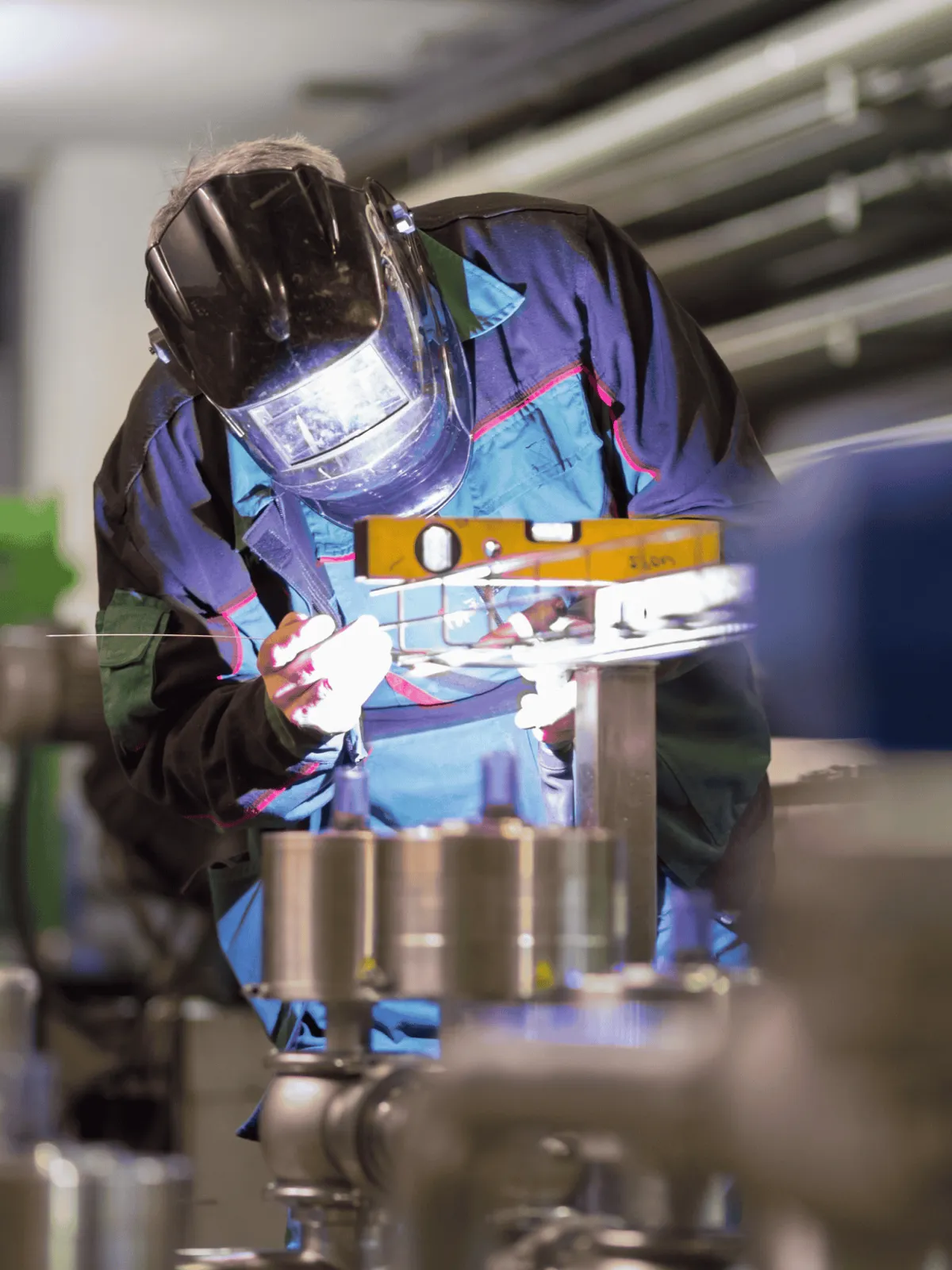 A worker in protective gear welding metal in an industrial fabrication in West Mountain Utah setting, surrounded by tools and pipes.