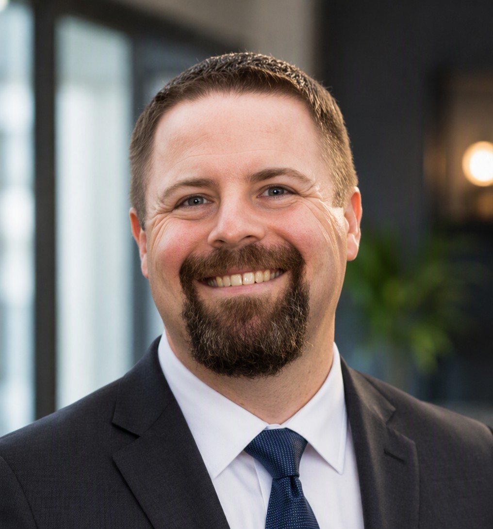 A professional headshot of a smiling Caucasian man with short brown hair and a prominent goatee. He is dressed in business attire, wearing a dark grey suit jacket, a crisp white collared shirt, and a dark blue patterned tie, set against a blurred office background.