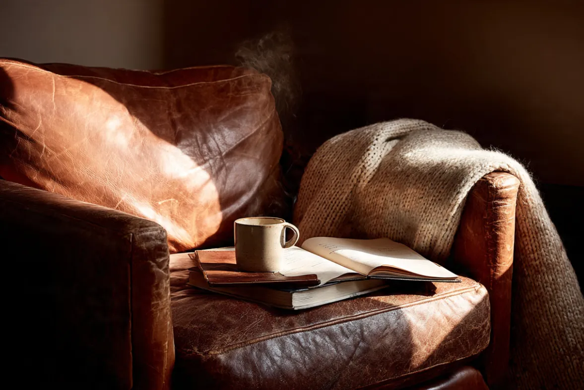 An intimate, eye-level shot of a well-worn leather armchair in a quiet corner. A soft, cream-colored blanket is draped casually over one arm. On the seat rests an open journal with handwritten notes and a steaming ceramic mug. A single beam of warm morning light cuts through the room, illuminating the steam rising from the mug. The walls are a soft, deep neutral tone. Lighting: Warm, directional morning light creating deep, comforting shadows. Mood: Sanctuary, quiet contemplation, the space where inner work happens.
