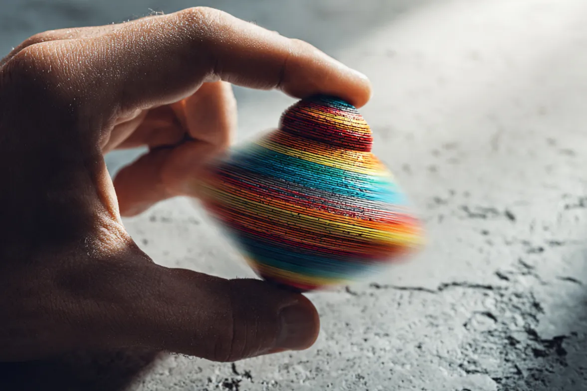 A close-up of a human hand holding a spinning top (like a dreidel or fidget spinner) with the six hat colors painted as stripes. The top is in motion, creating a vibrant blur of colors that symbolizes the dynamic process of switching perspectives. The background is a clean, textured concrete surface with subtle light rays (like sunlight through a window) casting soft shadows. The mood is energetic, modern, and slightly playful—like the mental agility of trying on new ways of thinking. 