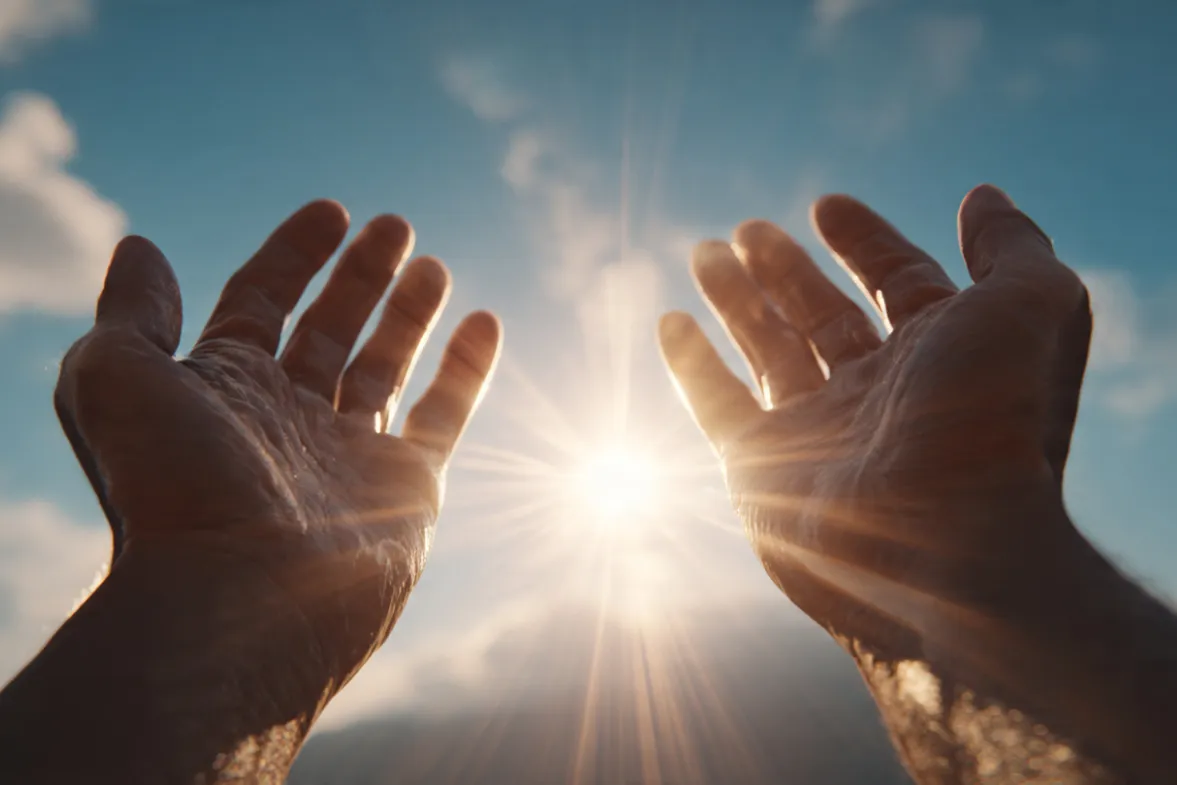 Close-up of open hands palms up against a brilliant blue sky with golden sunrise light streaming through fingers. Warm lens flare, soft clouds in background. No face visible—just the gesture of receiving.