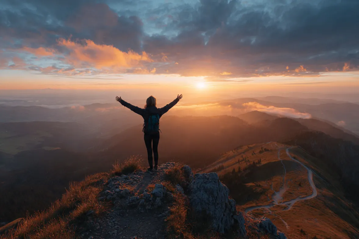 Woman with arms up celebrating the journey as the sun comes up, then she can see her the trail and how far she has come.
