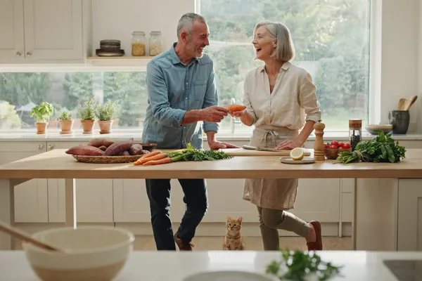 A vibrant, healthy, and stylish elderly couple in their 70s in a beautiful, sun-drenched modern kitchen. They are laughing joyfully while preparing a colorful, healthy meal together. On the counter, there are fresh sweet potatoes, colorful vegetables, and herbs. The man is handing a fresh ingredient to the woman. They look energetic, deeply connected, and full of life.
