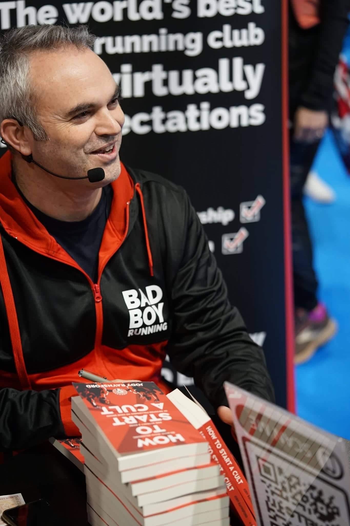 Portrait of Jody Raynsford, smiling, seated in a cozy book-lined office, surrounded by notes and a copy of 'How To Start A Cult', exuding approachability and expertise.