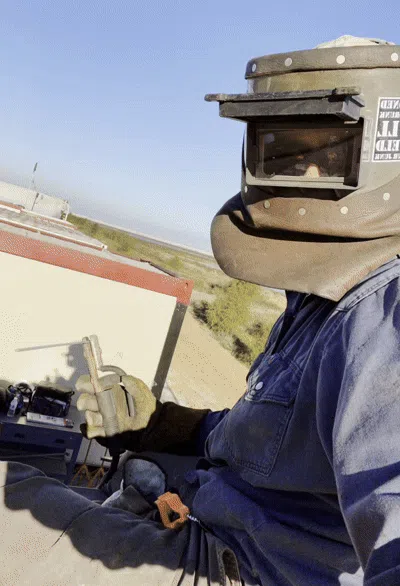 Welder in home garage, smiling, wearing safety gear, with a small welding table and tools in the background. Natural daylight, casual setting, middle-aged man, approachable demeanor.