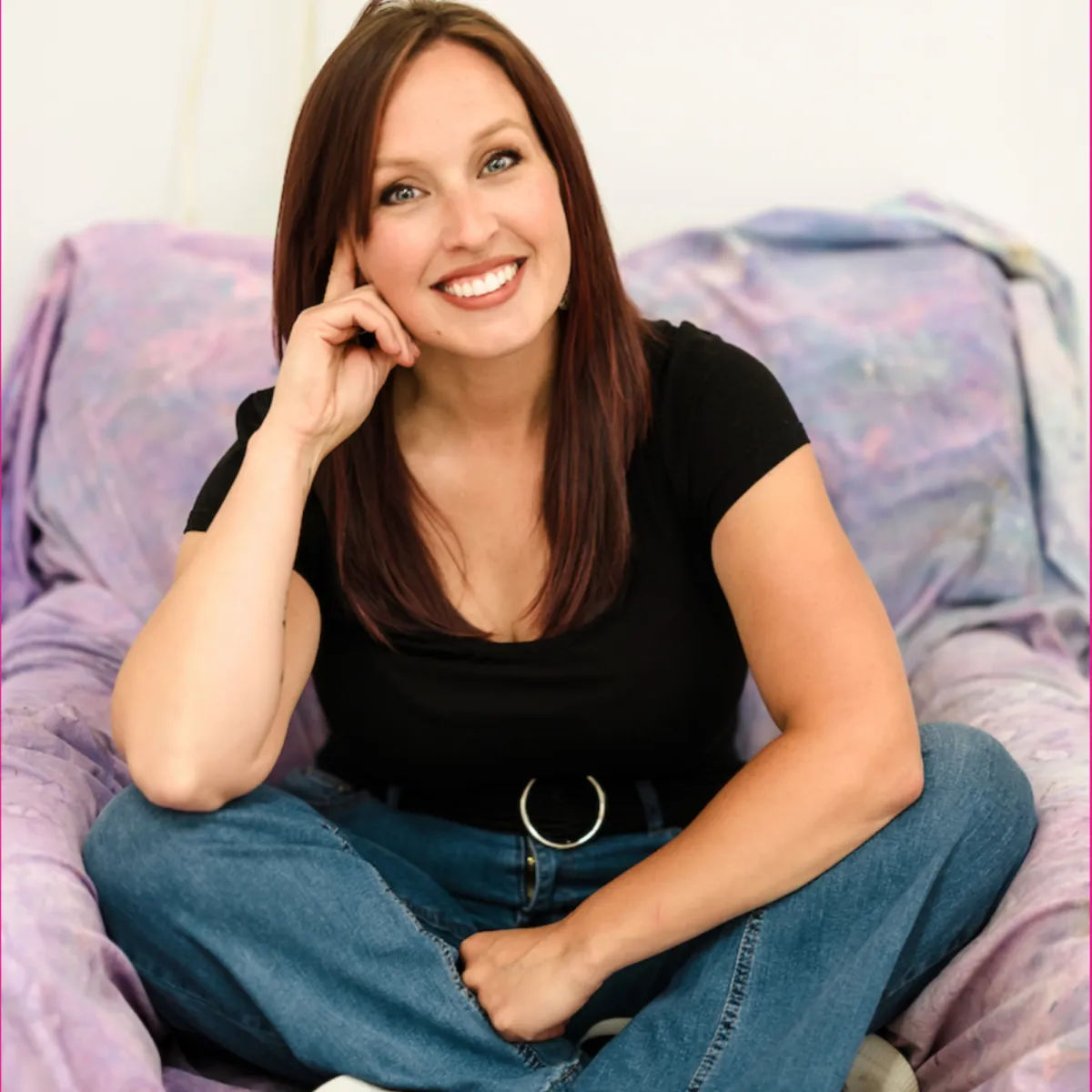 woman with long brown hair wearing black shirt and jeans sitting on an armchair draped in sheet splattered with colors
