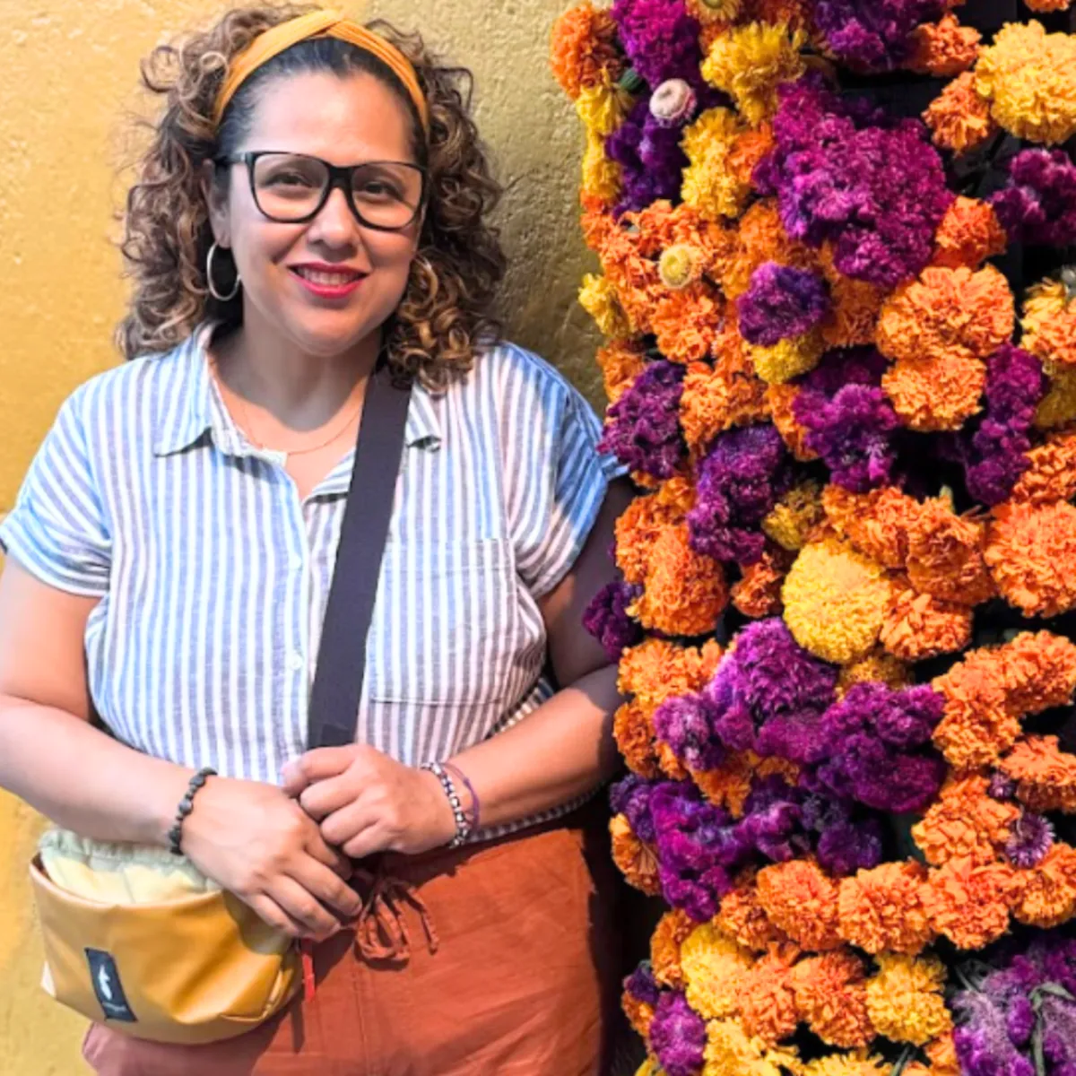 woman with curly hair standing against yellow wall wearing striped shirt and orange pants surround by flowers