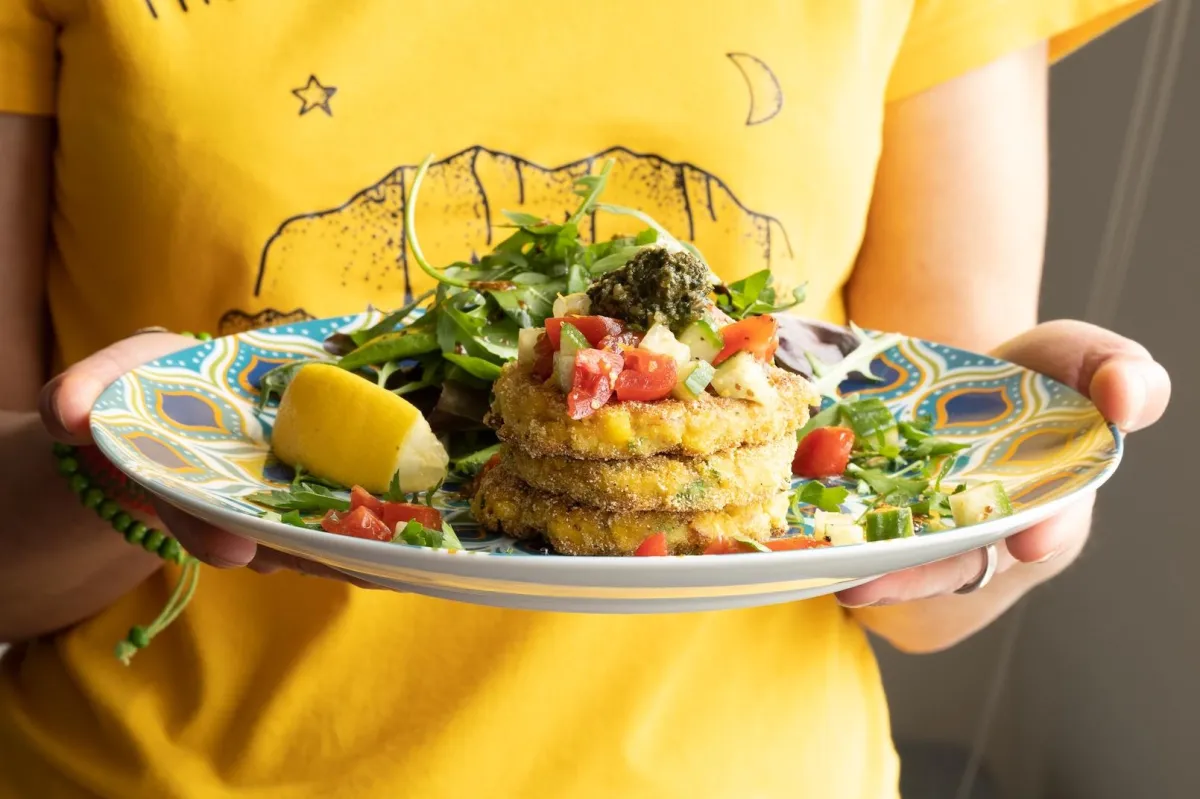 A woman holding a plate of sweecorn fritters topped with fresh pesto, with a lemon wedge and salad; homemade whole food suitable for vegans