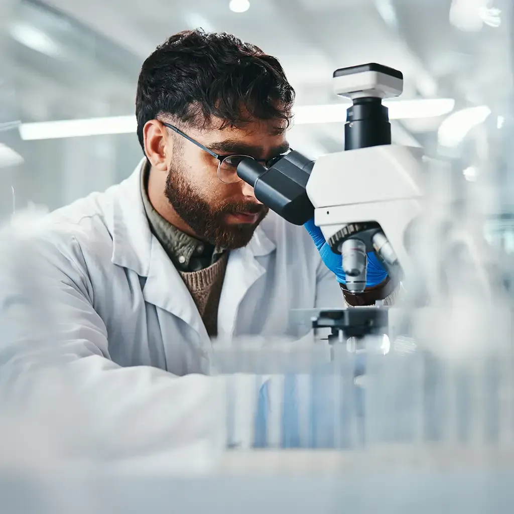 A man in a lab coat examines a specimen through a microscope in a laboratory setting.
