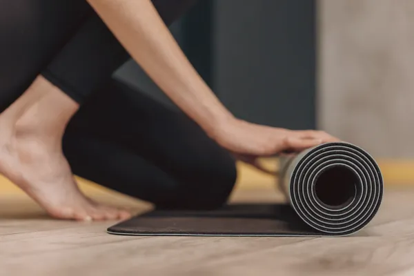Close-up of a person's hands rolling out a dark yoga mat on a wooden floor