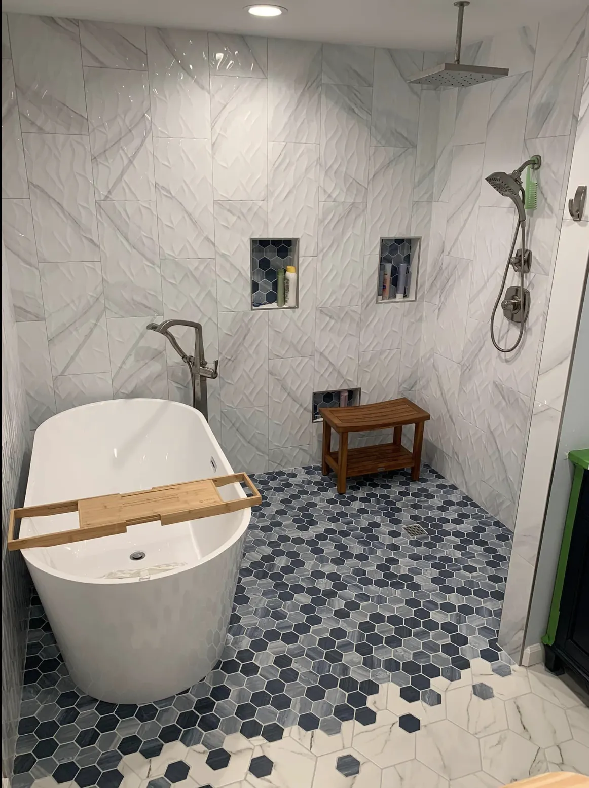 Luxurious bathroom with a freestanding soaking tub and modern rainfall shower. Features white textured wall tiles, blue hexagonal floor tiles blending with white, recessed shelves with hexagonal tile accents, and a wooden shower stool for a spa-like ambiance.