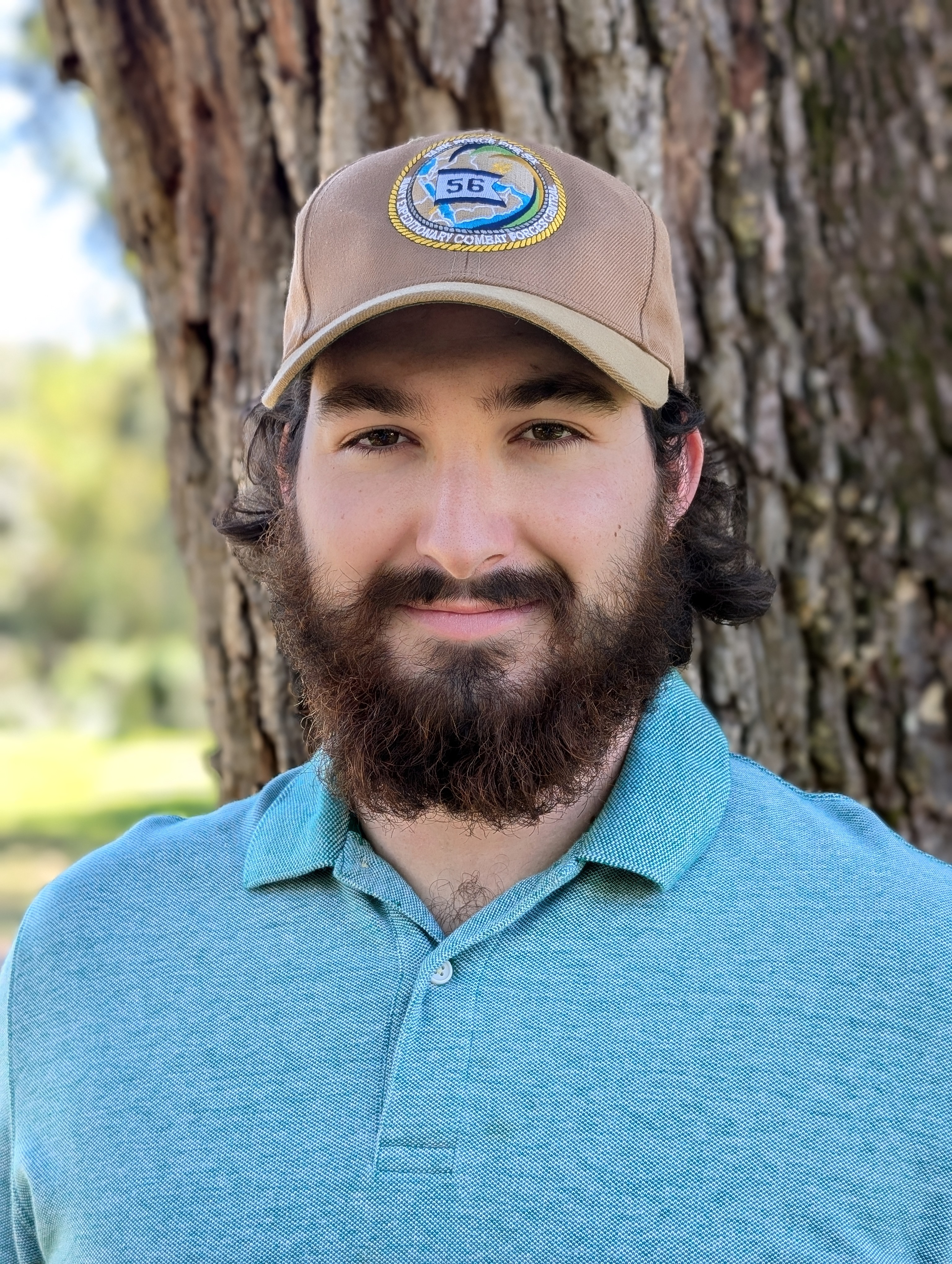 Headshot of a team member from The Custom Shower Company wearing a green polo shirt and a tan hat with a patch logo, smiling with a friendly demeanor in front of a tree, reflecting professionalism and warmth.