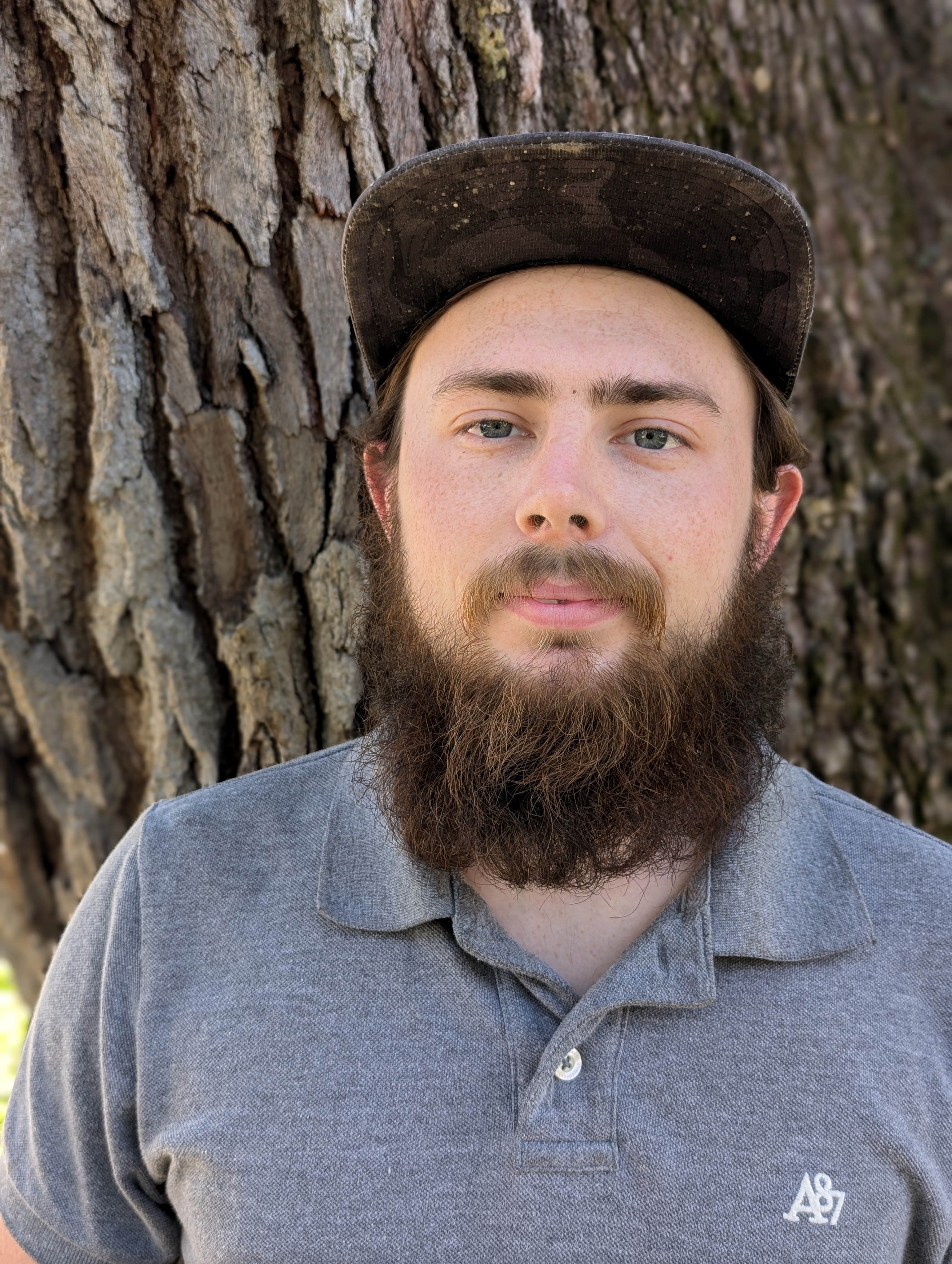 Headshot of a bearded team member from The Custom Shower Company wearing a gray polo and a baseball cap, standing in front of a tree, conveying a relaxed and approachable personality.