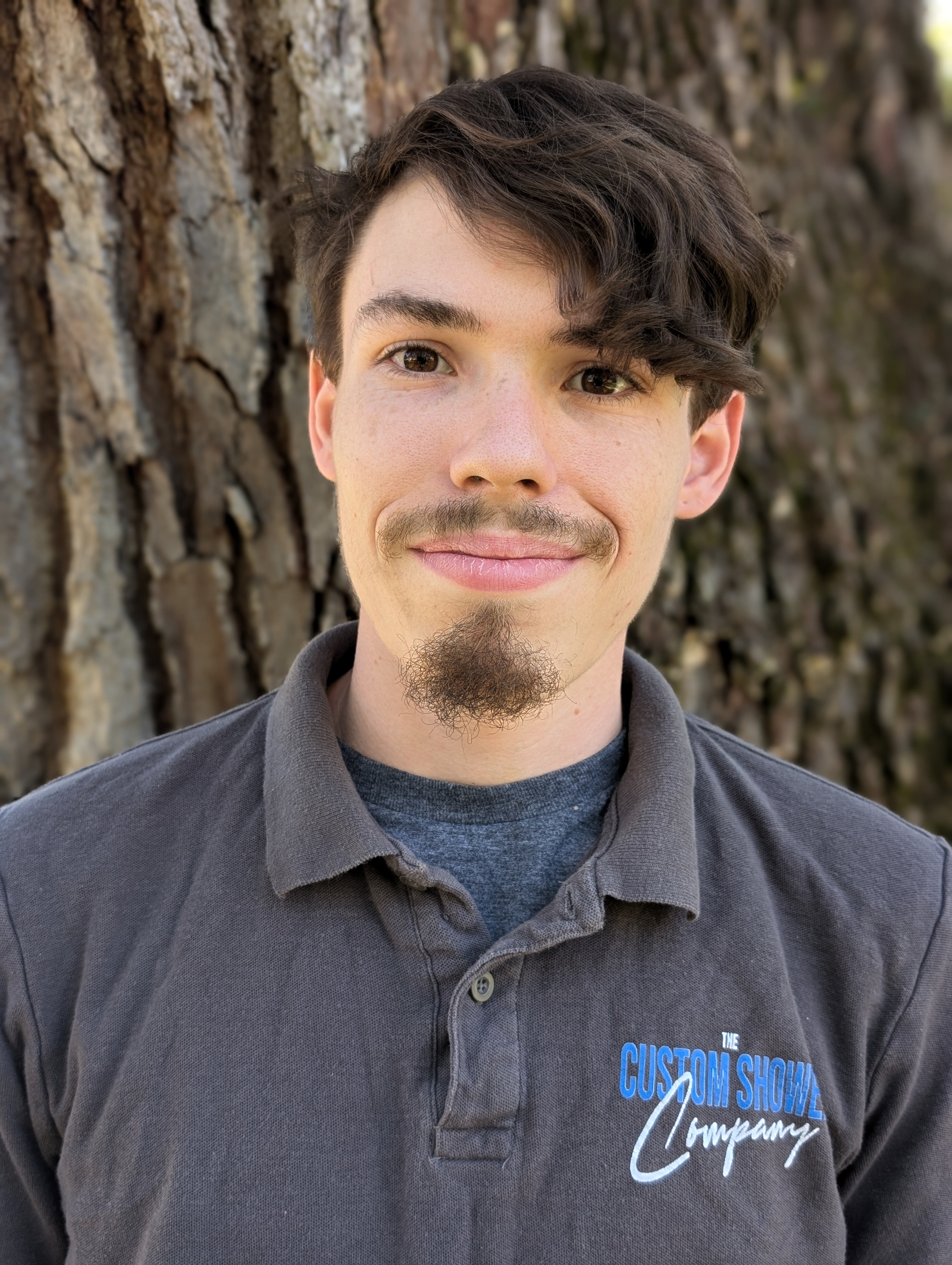 Headshot of a team member from The Custom Shower Company wearing a gray polo shirt with the company logo, smiling warmly against a tree background, representing professionalism and dedication.