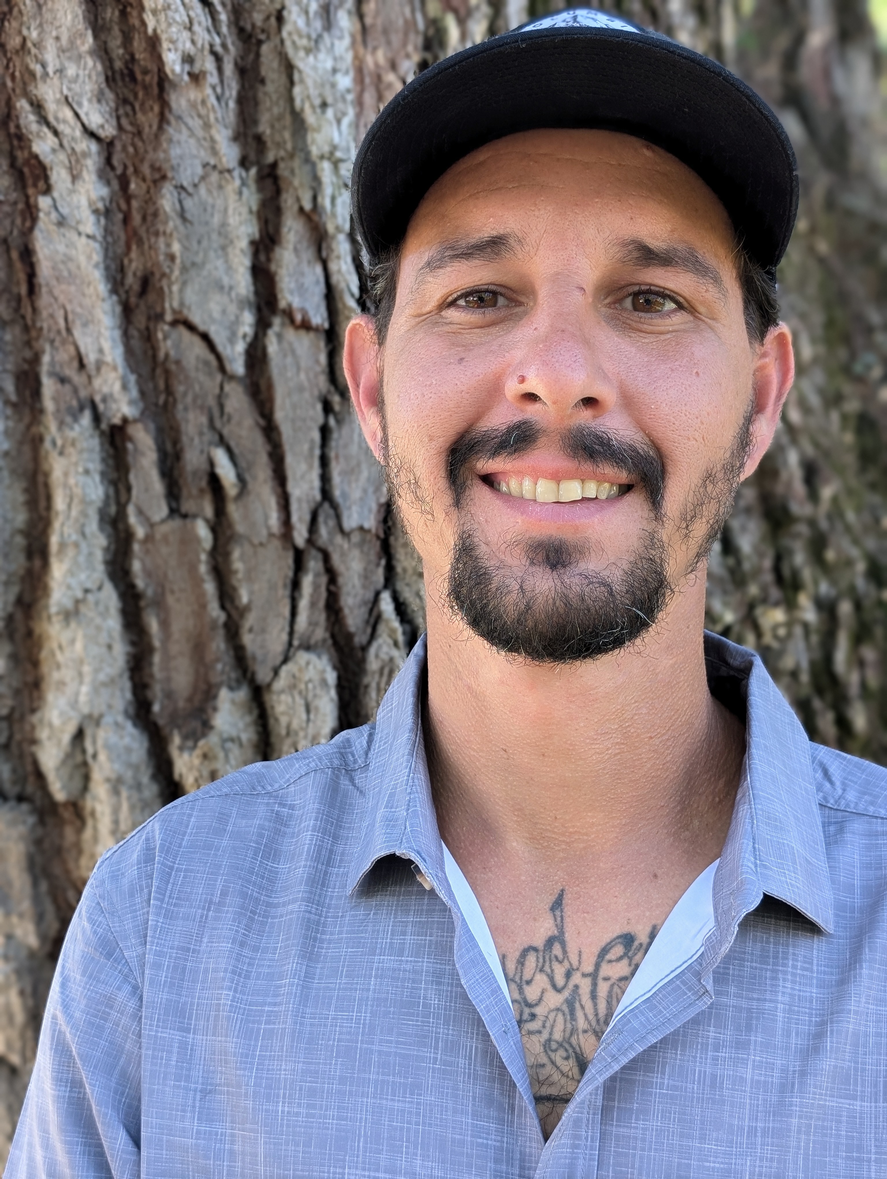 Headshot of a company founder of The Custom Shower Company with a goatee and wearing a gray button-down shirt and a black cap, smiling confidently with a tree trunk background, symbolizing experience and reliability.