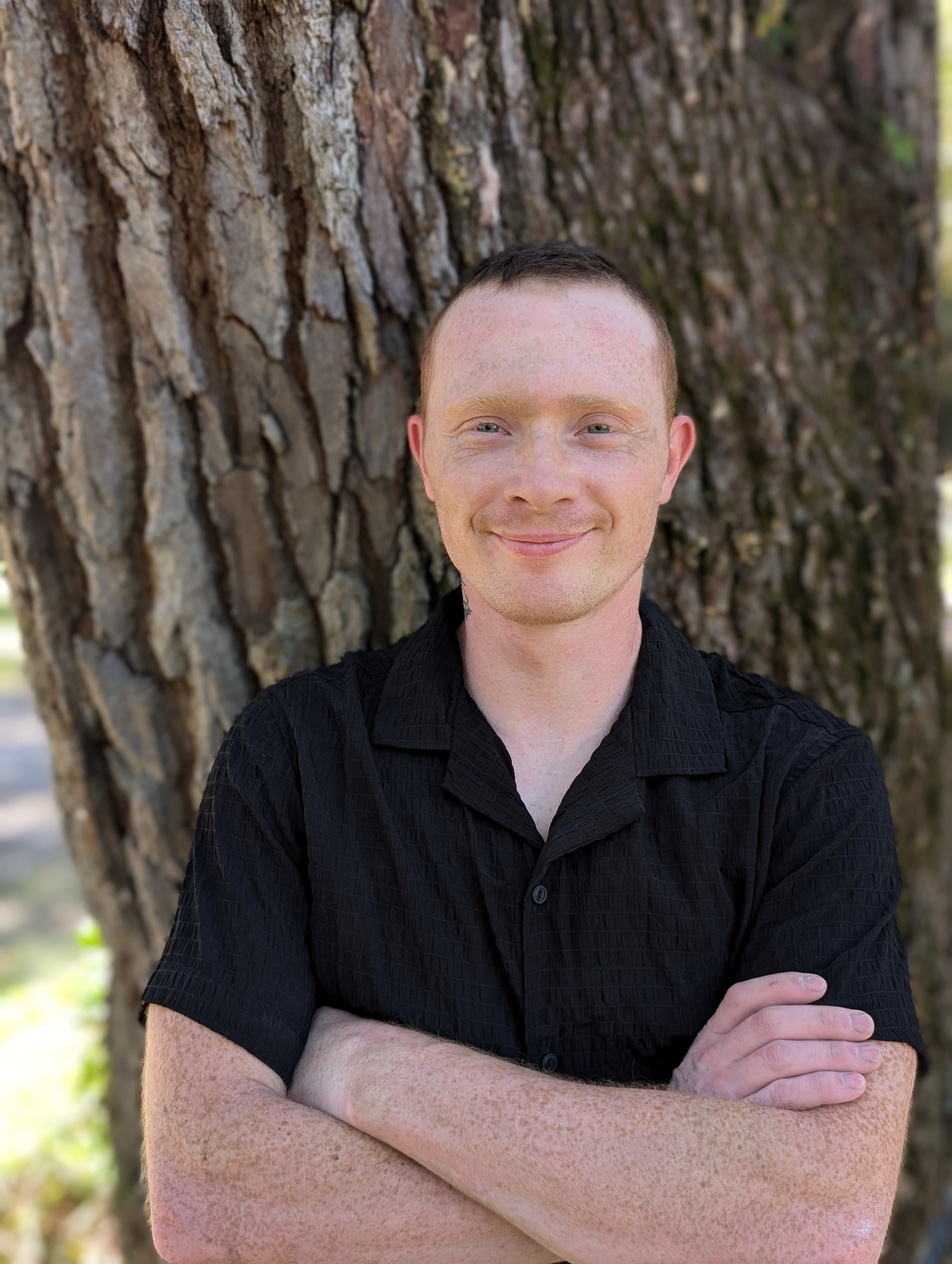 Headshot of a team member from The Custom Shower Company, smiling with arms crossed, wearing a black shirt and standing in front of a tree, creating a friendly and approachable impression.