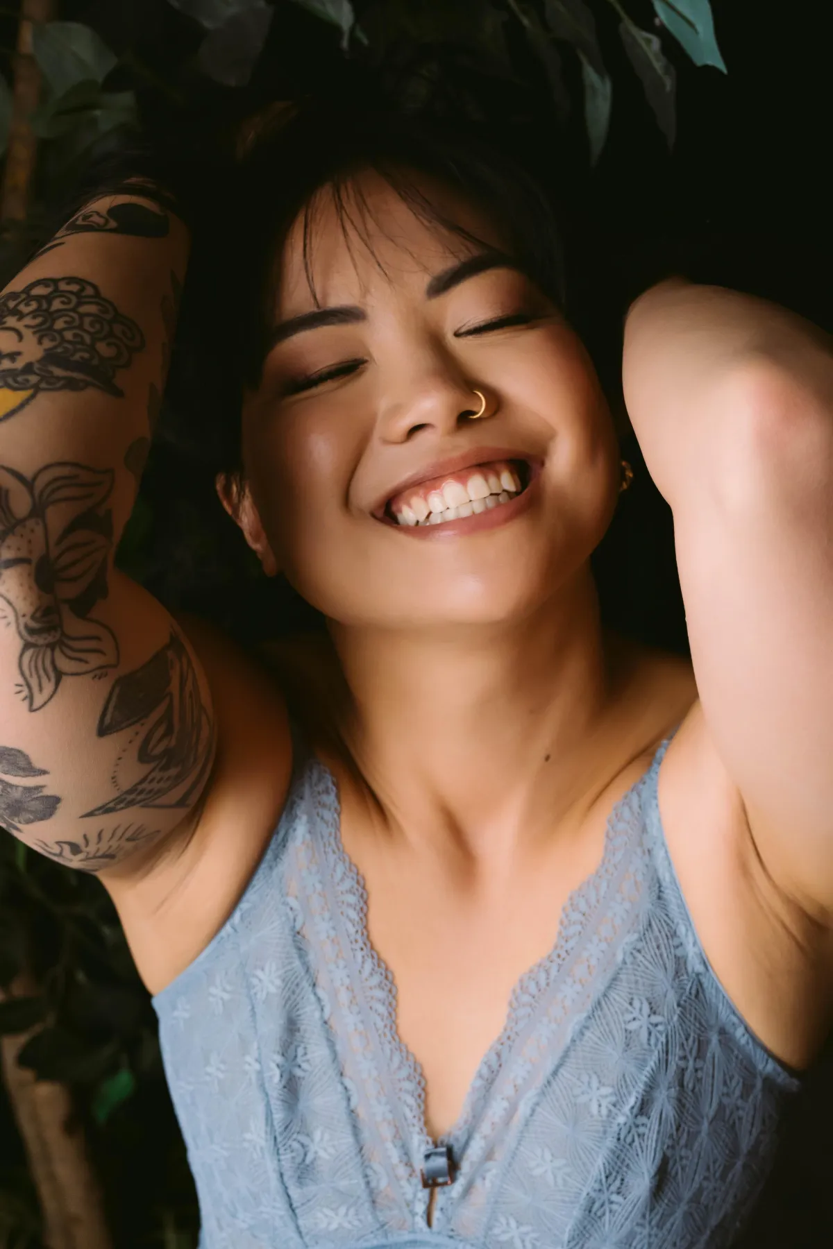 A black haired Asian woman with traditional black and white tattoos on one arm has her head reclined in laughter while wearing light, blue lacy lingerie.