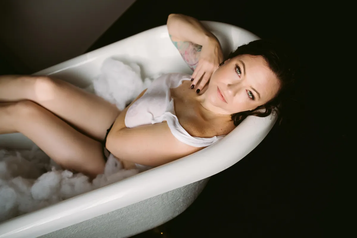 A woman with black hair and green eyes in a wet tank top reclines in a ceramic claw foot bath tub surrounded by bubbles. 
