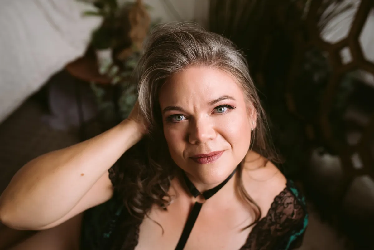 A woman with brown and grey hair with green or grey eyes looks up at the camera with a confident, soft smile. She is wearing a harness and a green and black lace lingerie bodysuit.