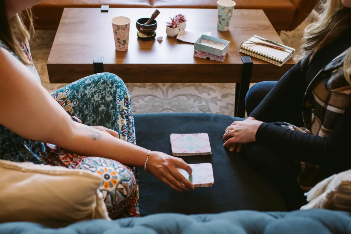 A woman in a blue patterned dress picks a oracle card from a stack of cards on a navy couch while she is sitting next to another woman in a black long sleeve shirt with a patterned vest.