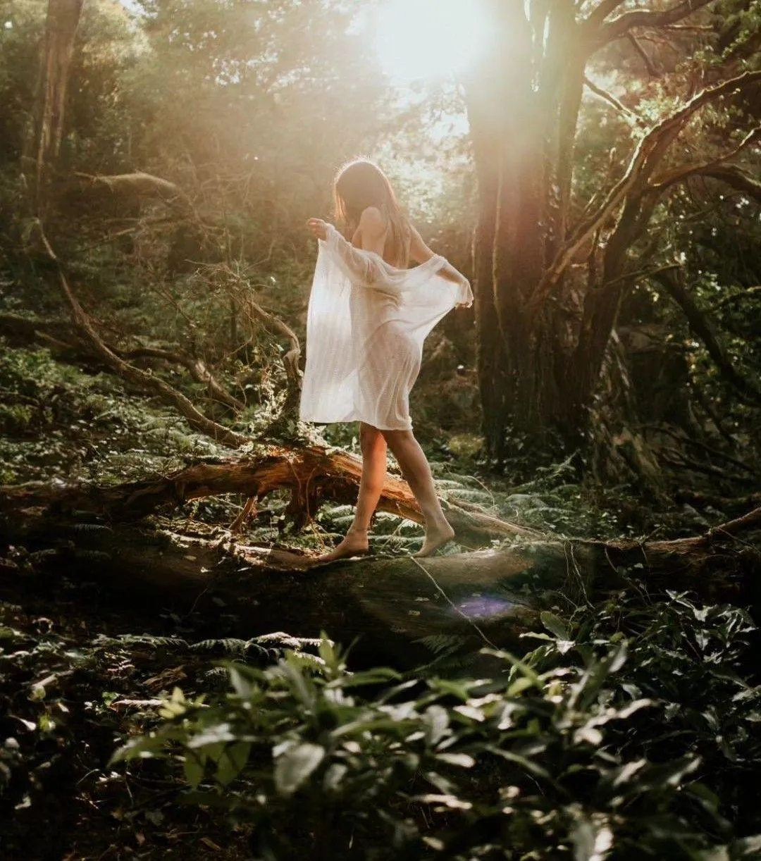 A brunette woman in a sheer white shirt walks on a log in the forest under the sunshine.