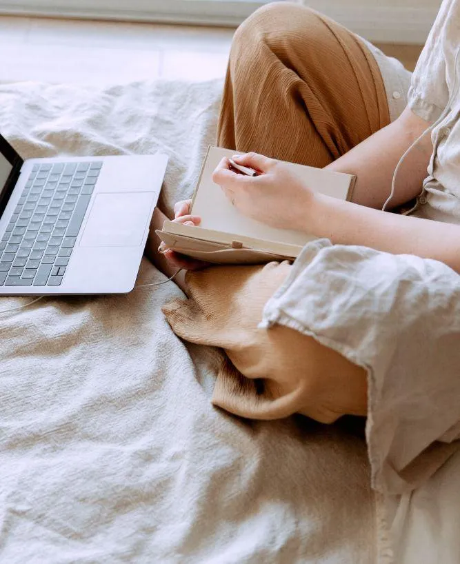 Woman sitting crosselegged on a bed with laptop in front of her, writing in notebook, with corded headphones