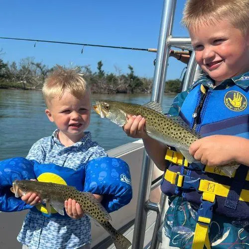 “Two smiling children proudly holding speckled trout during a family-friendly fishing charter with Captain Tanner in Gulf Shores, Alabama.”