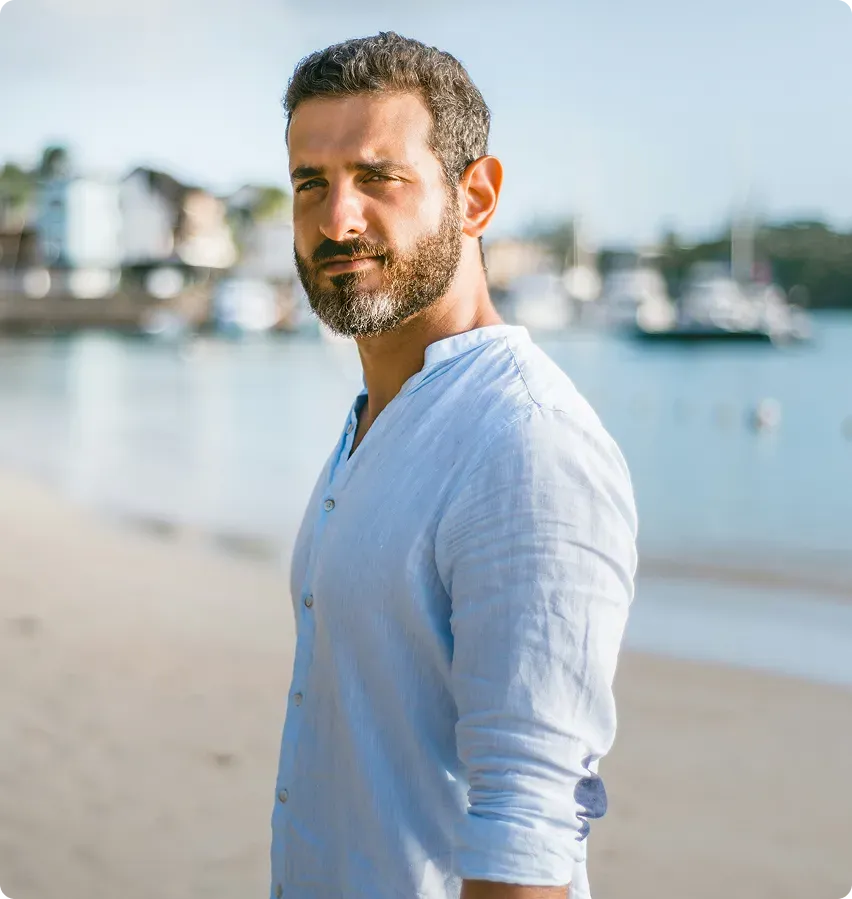 Confident man standing on the beach in a light blue shirt, symbolizing strength, calmness, and balanced masculine energy.