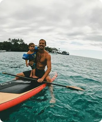  Man paddling on a surfboard with a young child sitting on the board, both smiling and enjoying a calm turquoise sea under a cloudy sky near the coast.