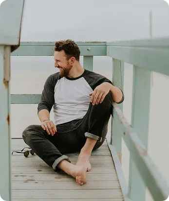 Man sitting barefoot on a wooden beach lookout deck, smiling and looking to the side while dressed casually in a black-and-white raglan shirt and jeans.
