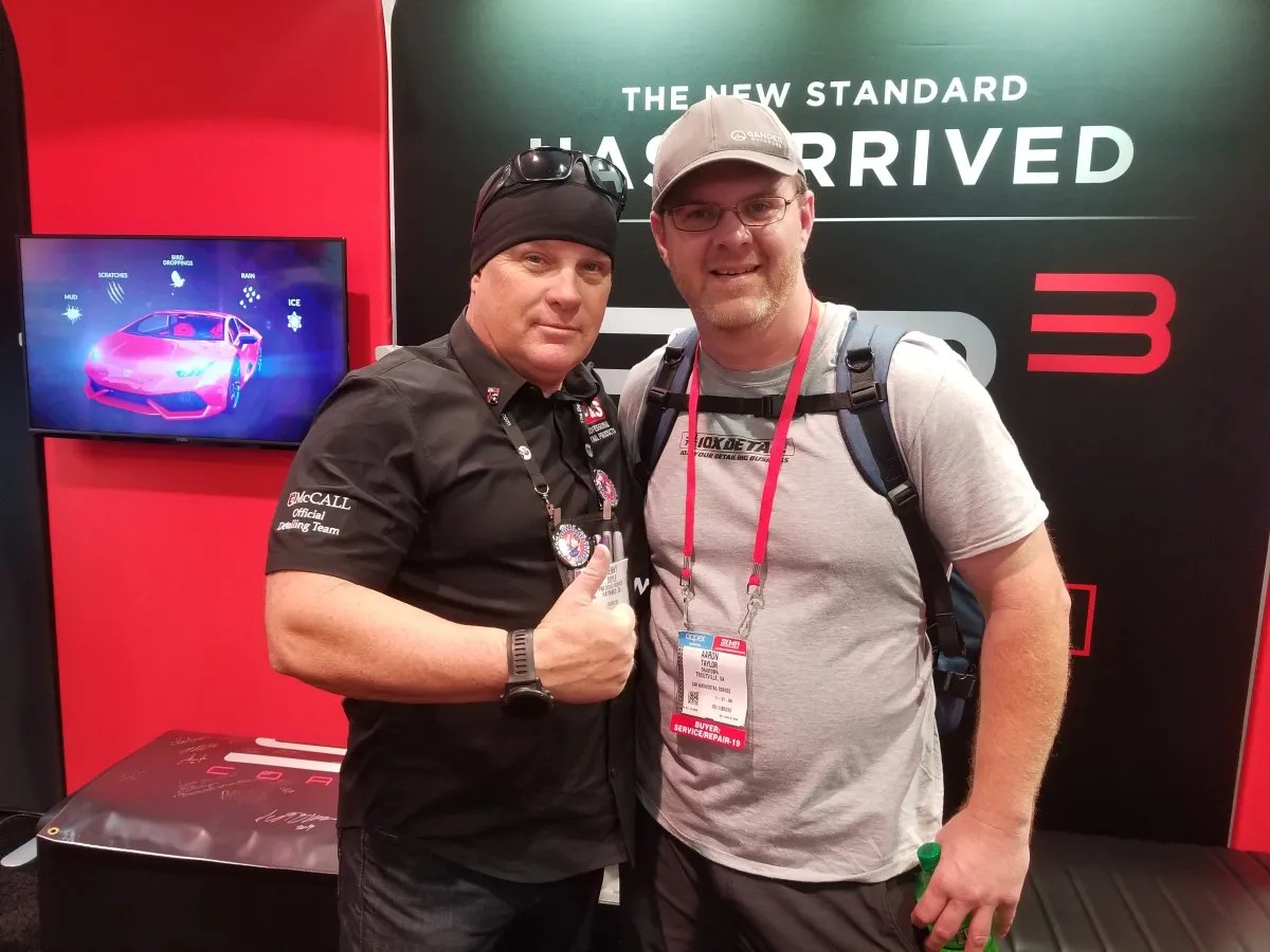 Two men at a trade show with a sign for "The New Standard Has Arrived," representing a paint correction Roanoke service.