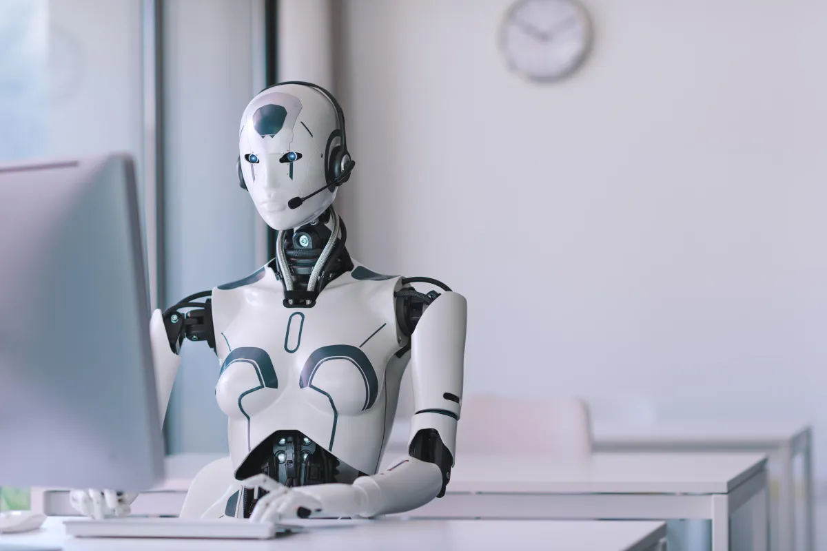 Picture of robot sitting at desk typing on computer with headset on talking to people