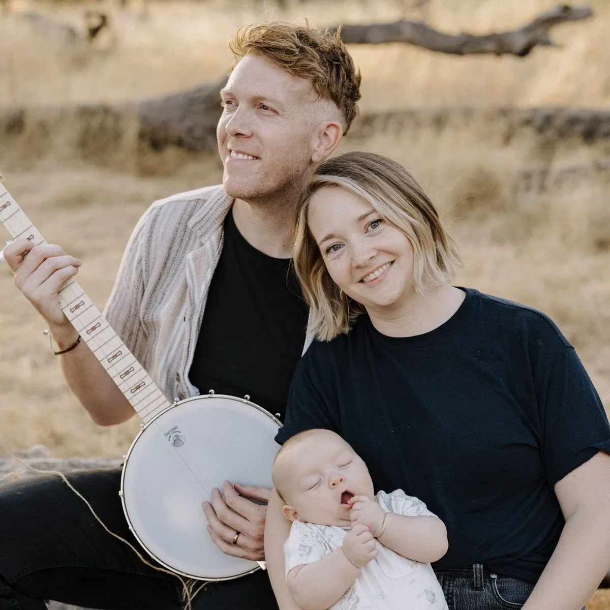 Smiling couple outdoors, the man holding a banjo and the woman holding their baby