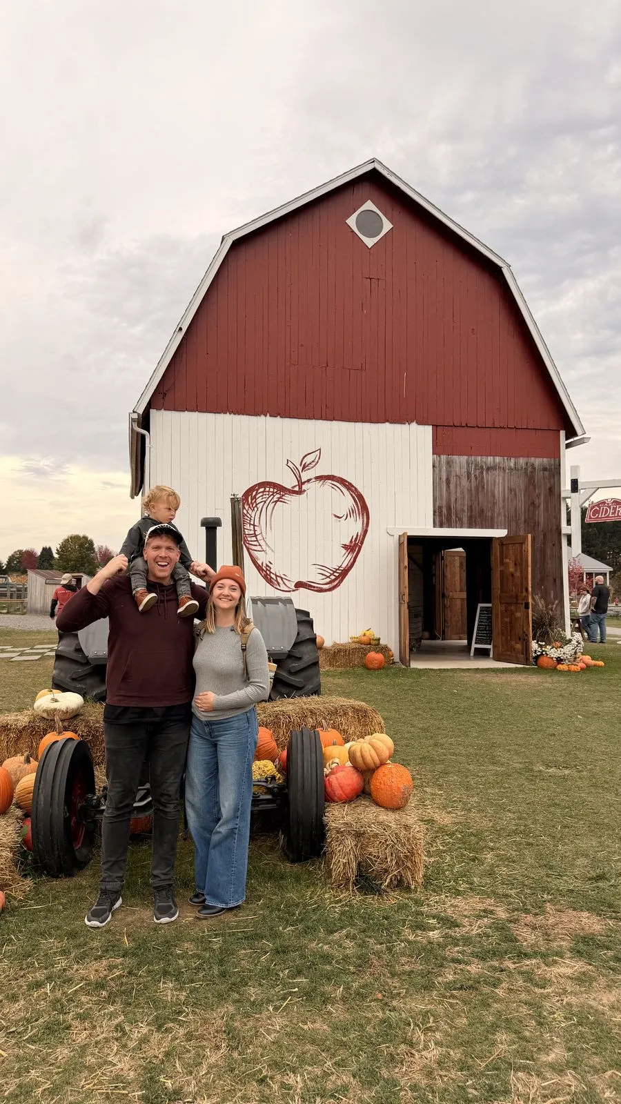 Family of four posing in front of a red barn with an apple painted on it, surrounded by pumpkins and hay bales