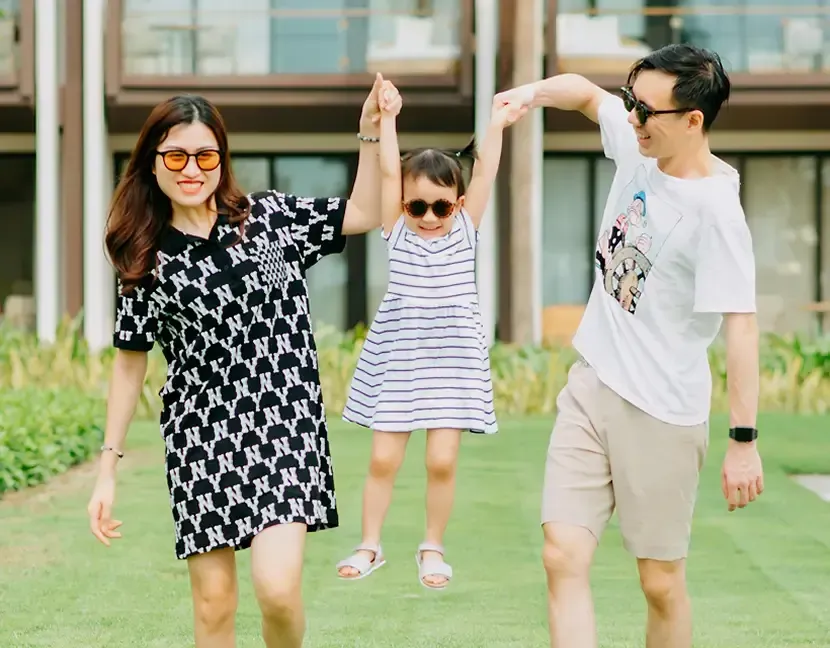 A man and a woman hold up a smiling child by the hands while walking on grass. All are wearing sunglasses. There are modern buildings and palm trees in the background.
