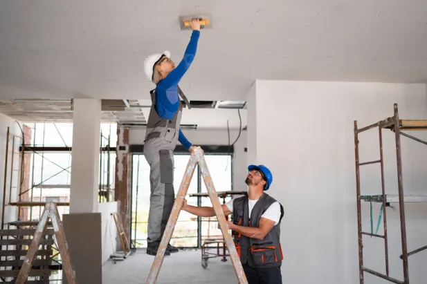 Two men installing ceiling panels in a room, using tools and wearing safety gear.