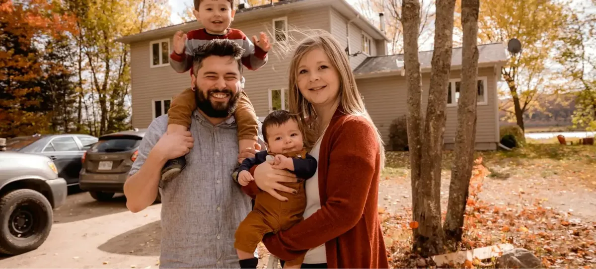 Four people sit on the front steps of a brick house, posing for a photo. Three adults wear casual winter attire, while a child in a red sweater sits between two adults.