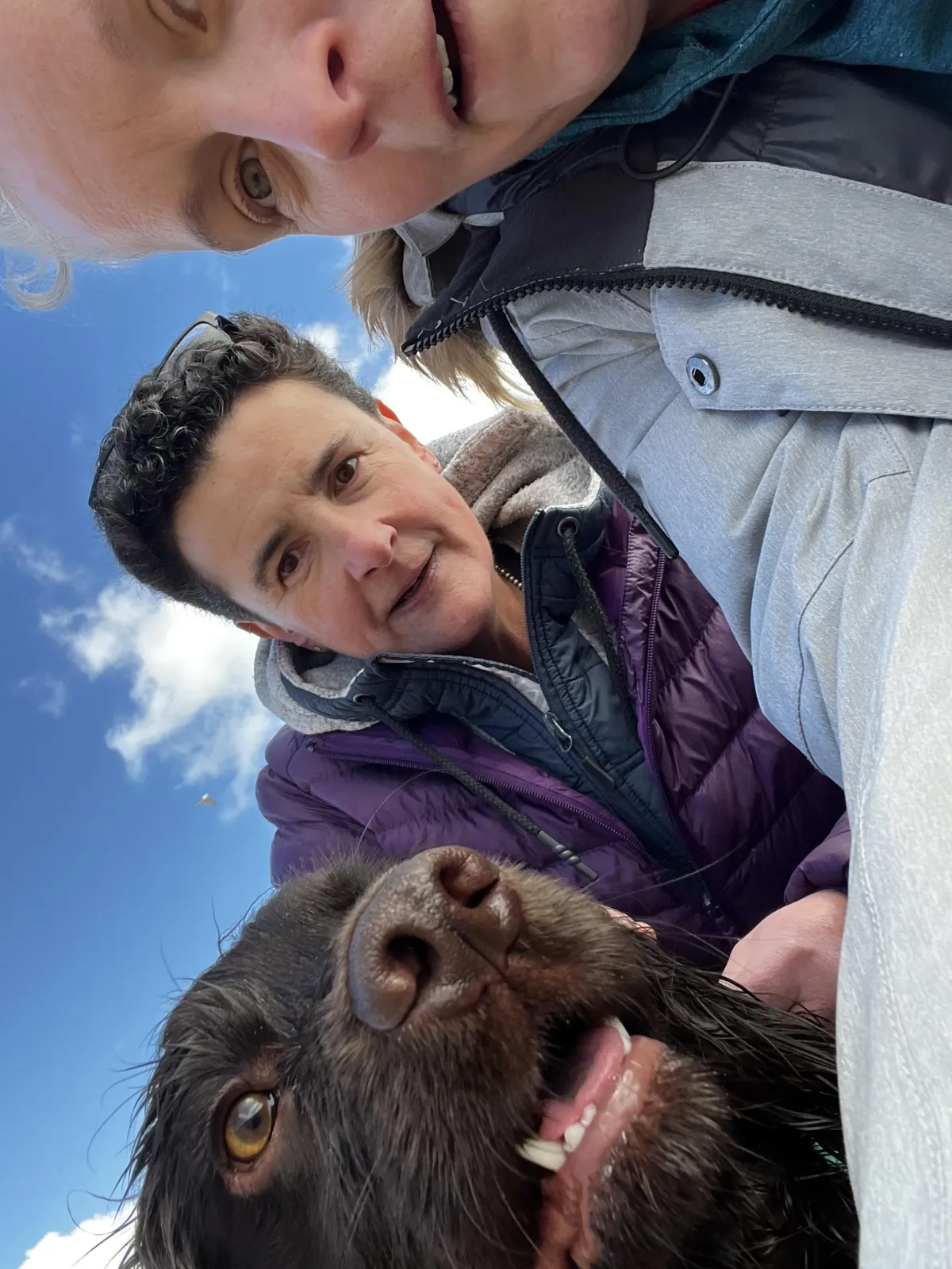 Sarah Watts with her wife and their cocker spaniel Kraken, smiling into the camera on a bright day outdoors — a joyful family moment full of love and muddy paws.