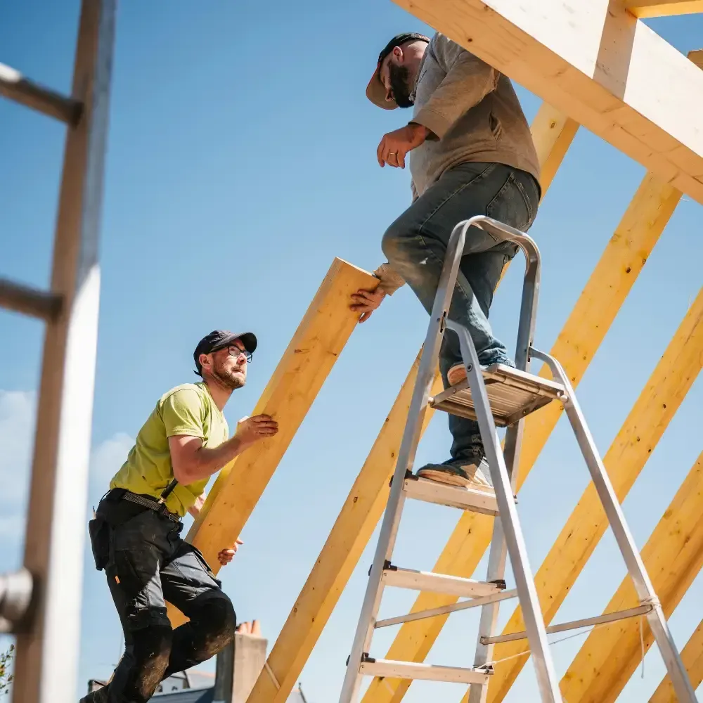 Two construction workers assemble a wooden frame outdoors under a clear blue sky. One worker stands on a ladder positioning a large wooden beam while the other, standing on the ground, steadies the beam and looks up to assist. The structure appears to be part of a roof frame under construction.