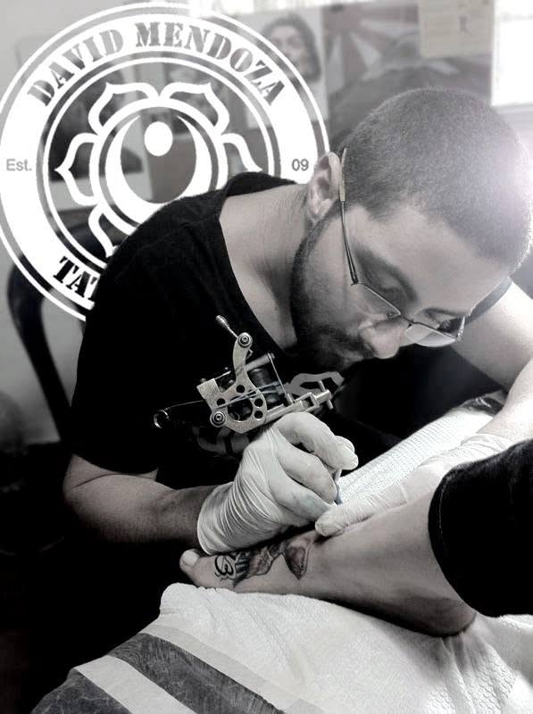 David Mendoza en sus inicios como artista del tatuaje. Esta foto el 2010, mientras tatúa a un cliente con su primer logotipo de fondo.