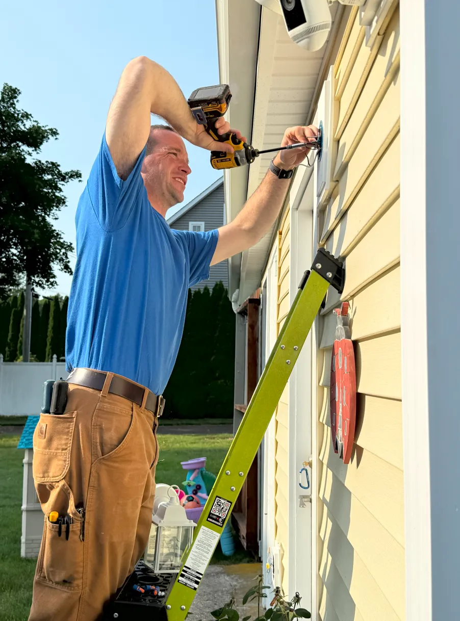Licensed electrician in Old Saybrook, CT installing a residential security camera system on a home’s exterior siding. Expert in residential and commercial electrical services along the Connecticut shoreline. Professional electrical contractor using power drill while standing on ladder for outdoor security installation.