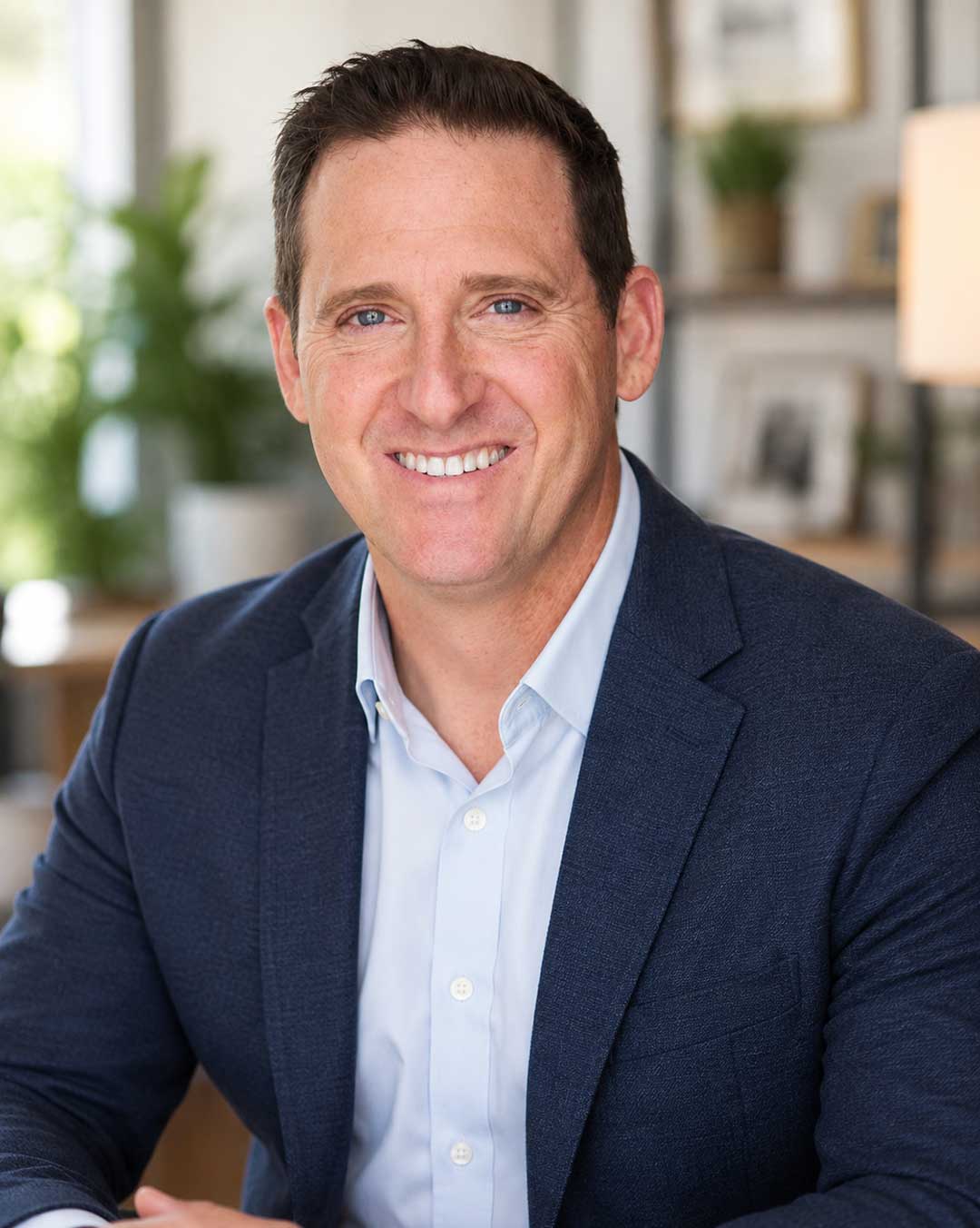 Professional headshot of a smiling man wearing a blue blazer and light blue shirt against a white brick wall.
