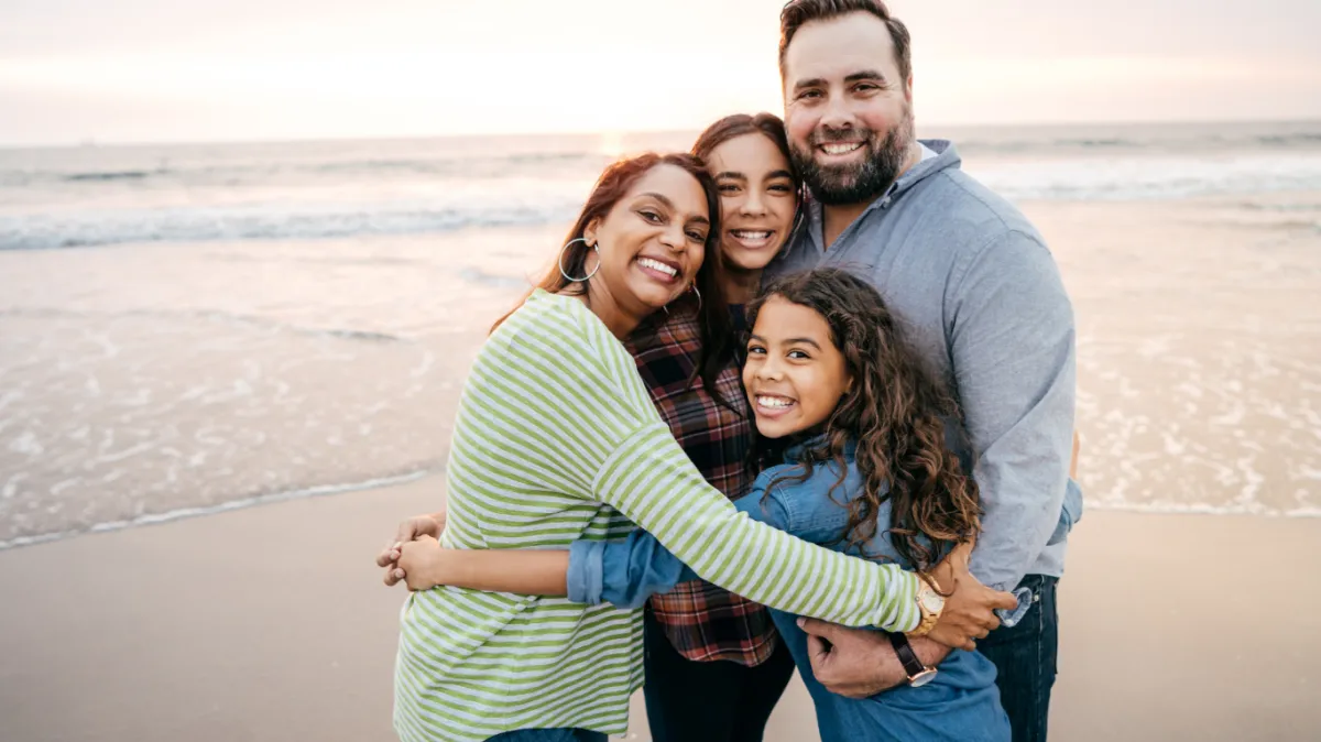 A smiling parent and their gender creative child sitting together on a sunlit living room couch, both looking at each other with warmth and trust. The parent gently holds the child's hand, conveying support and affirmation. The background features soft, pastel decor and a window with natural light.