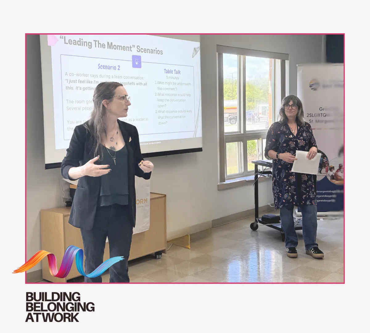 Two women present in a bright office classroom, one speaking in front of a whiteboard titled “Leading The Moment’ Securities,” while the other stands near a banner and holds papers.