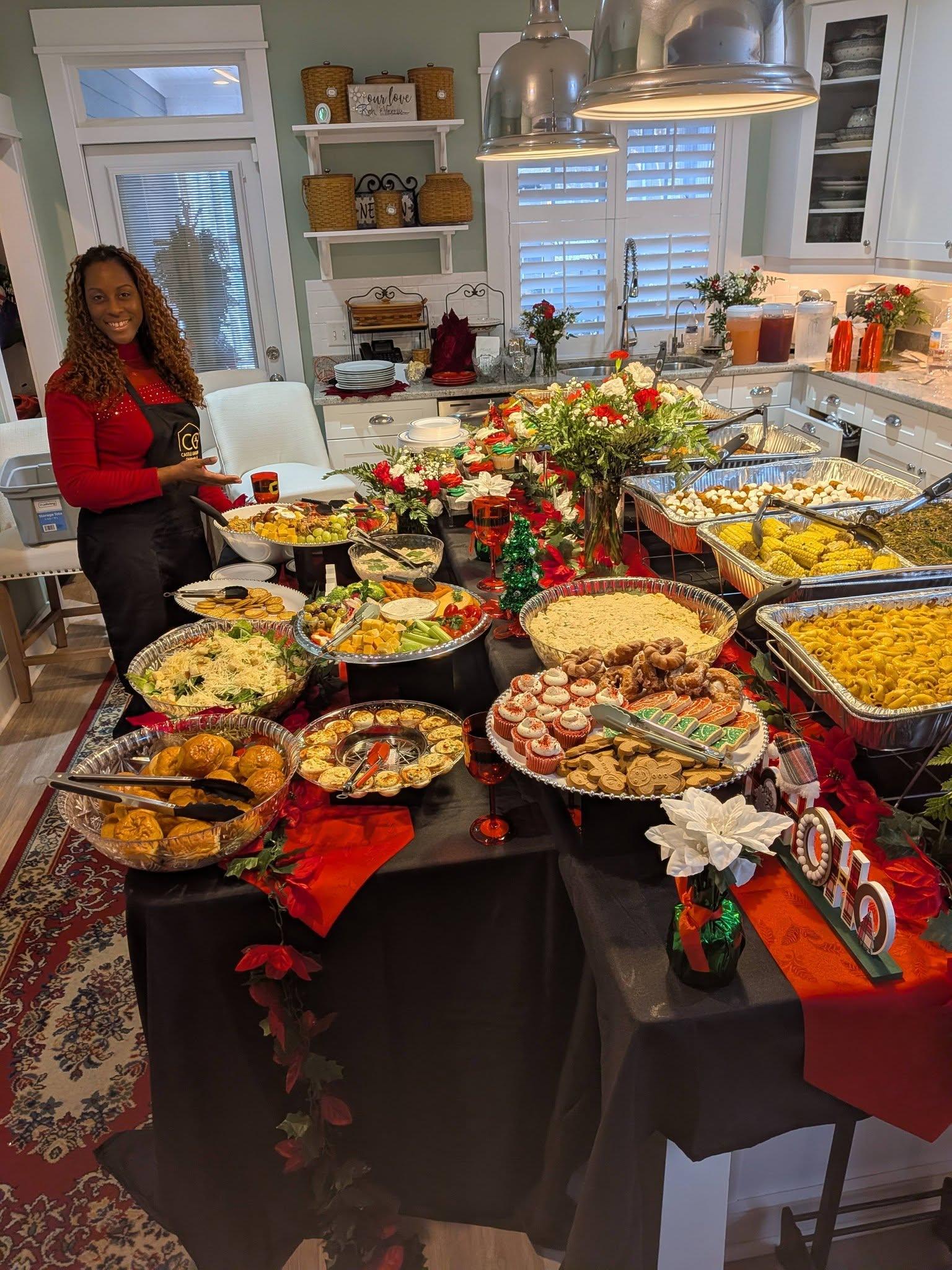 Buffet table adorned with festive food displays, including appetizers, desserts, and floral arrangements, with a smiling woman in a red sweater and apron, highlighting hospitality for gatherings at Castle Gardens ILM.