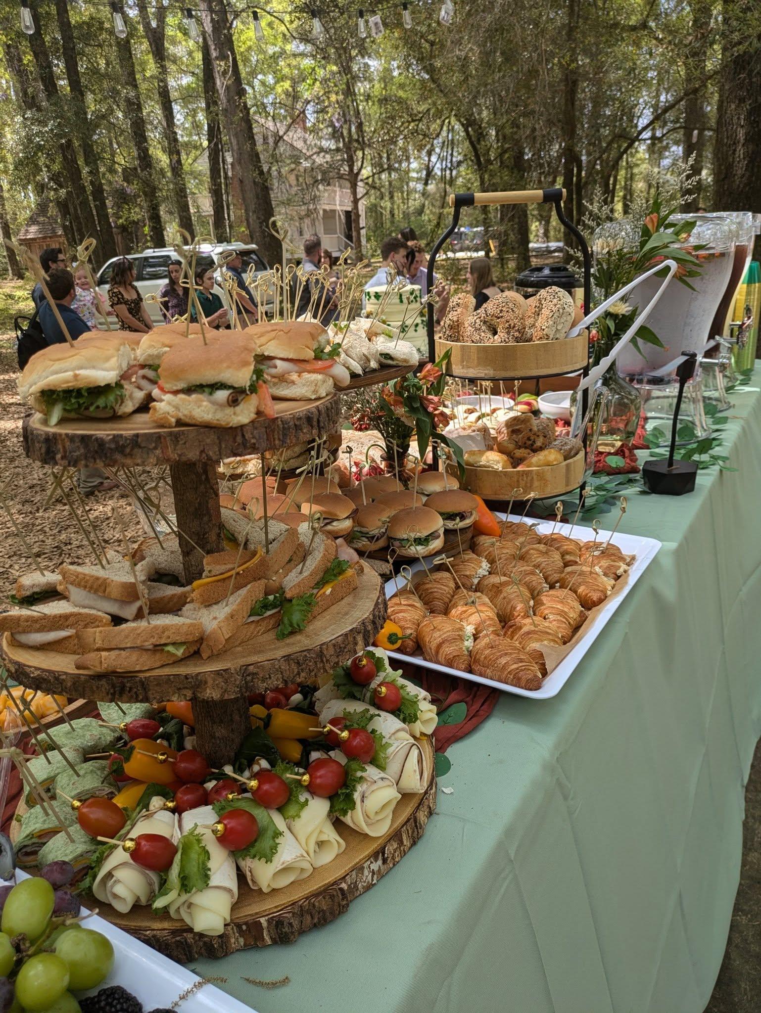 Elevated culinary display featuring assorted sandwiches, croissants, and vibrant vegetable garnishes, set in a natural outdoor venue for celebrations at Castle Gardens ILM.