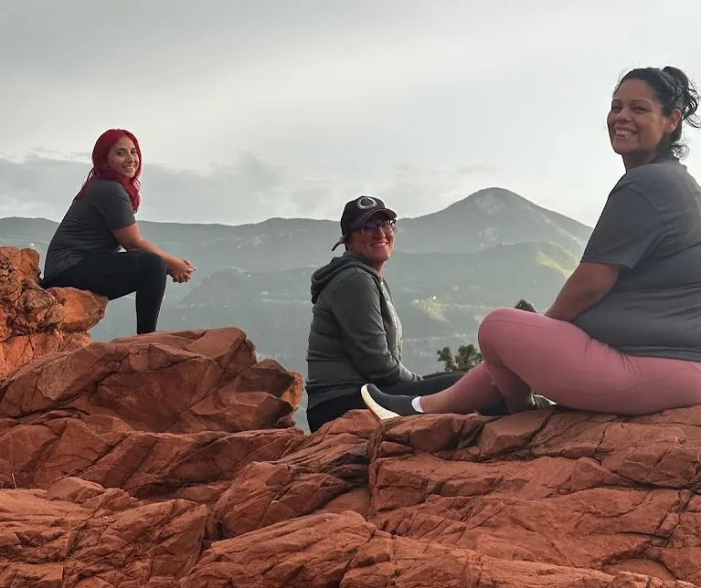 Three women sitting on red rock formations, smiling with mountains and a cloudy sky in the background.